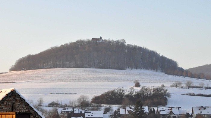 Verschneite ländliche Landschaft mit einer Burg auf einem Hügel und einem malerischen Dorf darunter bei klarem Himmel.