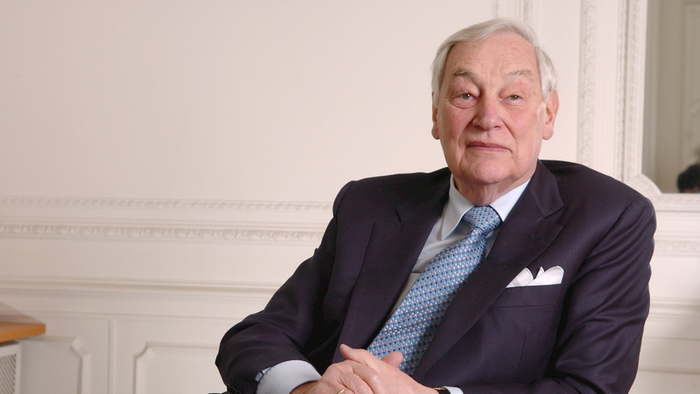 Ein älterer Mann im Anzug mit Krawatte sitzt nachdenklich an einem Schreibtisch mit Brille und Papieren.