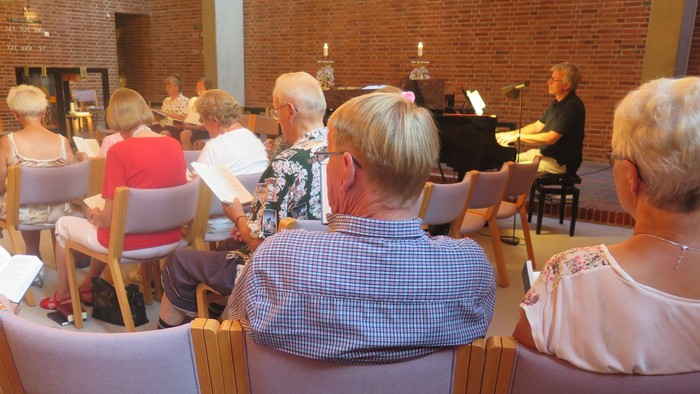 Gruppe af mennesker, der synger sammen i en kirke eller kapel med en pianist, der akkompagnerer dem.