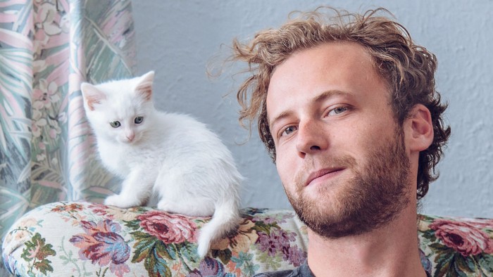 Junger Mann sitzt auf einem Sessel, neben ihm sitzt eine weiße Katze.