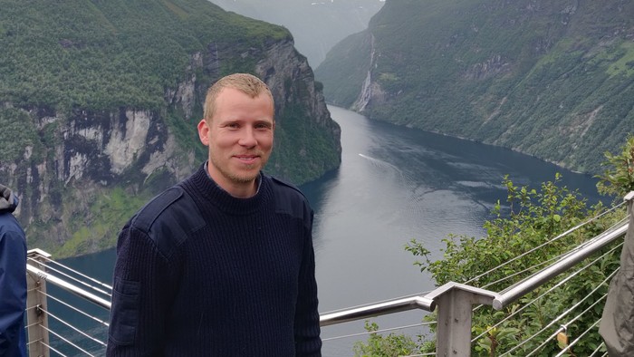 En person står og smiler ved en naturskøn fjord omgivet af stejle, frodige grønne klipper.