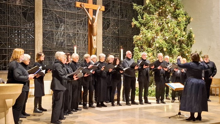 Ein gemischter Kammerchor in Schwarz singt im Halbkreis in einem modernen Altarraum. Im Hintergrund ein Weihnachtsbaum,