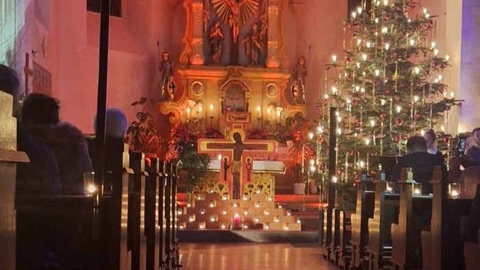 Auf dem Bild ist der mit Kerzenschein beleuchtete Altarraum der Kirche Hainzell zu sehen. In der Mitte steht das Taizé-Kreuz, erhellt durch viele Kerzen. Auf der rechten Seite ist ein prächtig geschmückter und leuchtender Tannenbaum zu sehen.