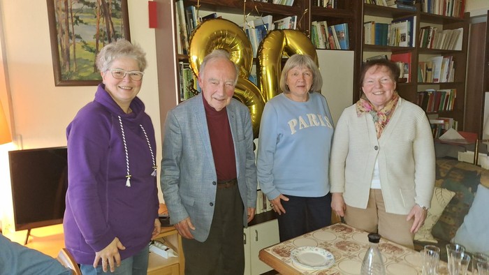 Vier Personen posieren gemeinsam drinnen mit einem goldenen Ballon in Form eines Saxophons und Regalen mit Büchern im Hintergrund.