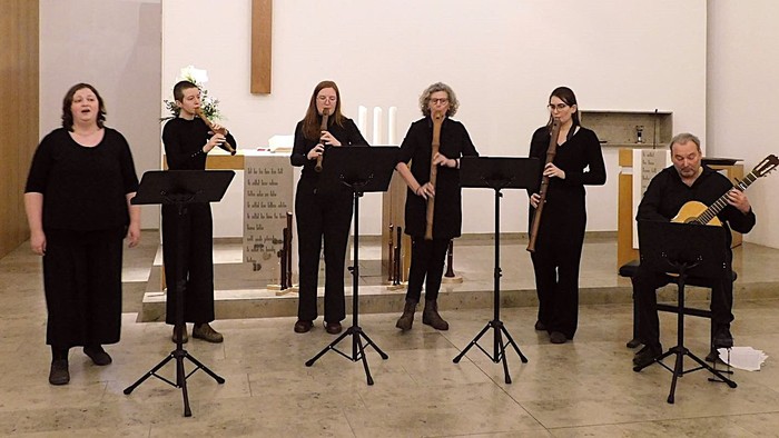 Eine Gruppe von Musiker:innen, die mit Holzblasinstrumenten, einer Gitarre und Sänger:innen in einer kirchenähnlichen Umgebung auftreten.