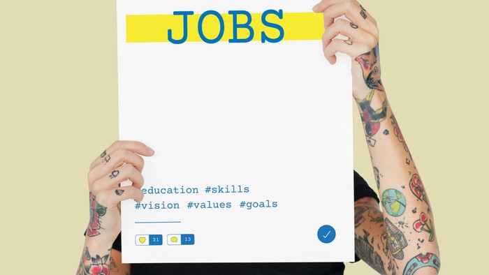 Person mit tätowierten Armen, die ein Schild mit der Aufschrift „JOBS“ hält, zusammen mit Hashtags zum Thema Karriereentwicklung.