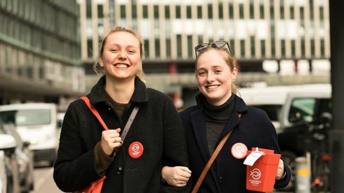 To glade personer går sammen og holder kaffekopper, mens de bærer tasker med røde logoer på en bygade.