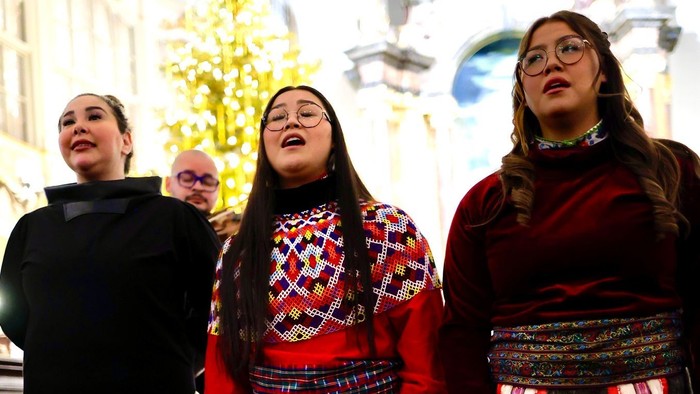 Tre kvinder i traditionelt farverigt strikket tøj, der synger sammen indendørs under en festlig begivenhed.
