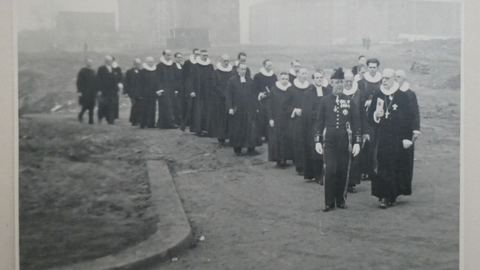 En sort-hvid procession af formelt klædte personer, der marcherer mod en kirke i højtidelig ceremoni.
