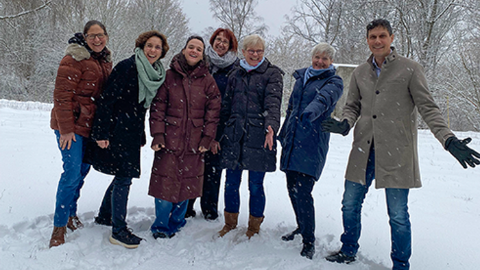 Sieben erwachsene Personen stehen gemeinsam im Freien auf einer verschneiten Fläche und blicken lächelnd in die Kamera. Alle tragen winterliche Kleidung wie Mäntel, Jacken, Schals und feste Schuhe. Leichter Schneefall ist sichtbar. Die Gruppe steht locker