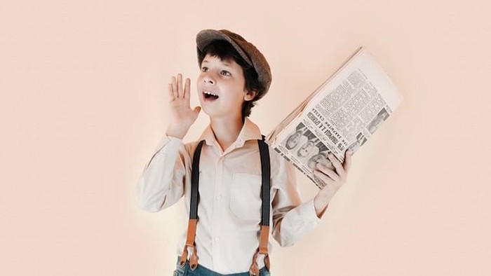 Ein Junge in Hemd, Hosenträgern und Hut hält eine Zeitung und zeigt überrascht nach oben.