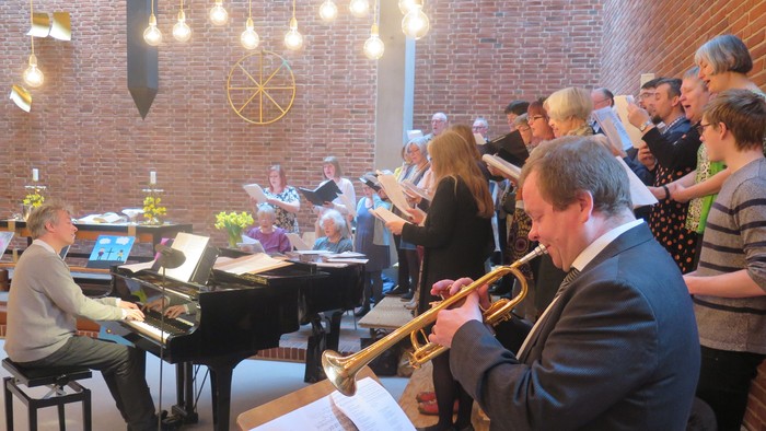 Et livligt kor og messingensemble optræder sammen i et varmt lokale med murstensvægge og noder.