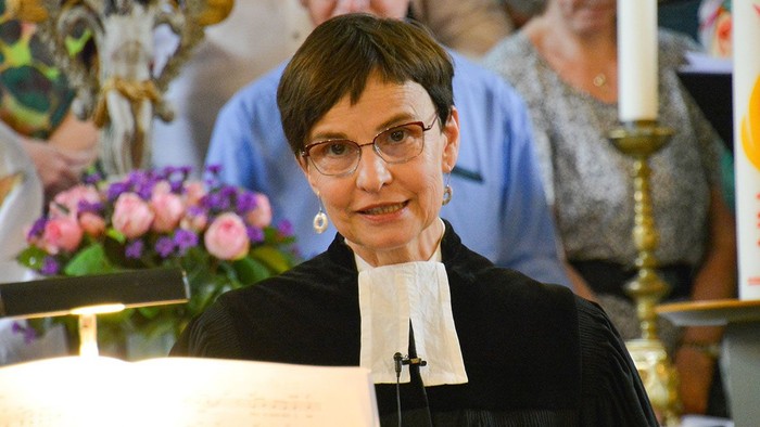 Pfarrerin in schwarzer Amtstracht mit weißem Beffchen steht am Lesepult in einer Kirche und spricht, im Hintergrund sind unscharf weitere Menschen sowie Kerzen und Blumenschmuck zu sehen.