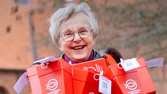 En smilende ældre kvinde holder flere røde indsamlingsbøsser og promoverer et fællesskabsindsamlingsarrangement den 8. marts.
