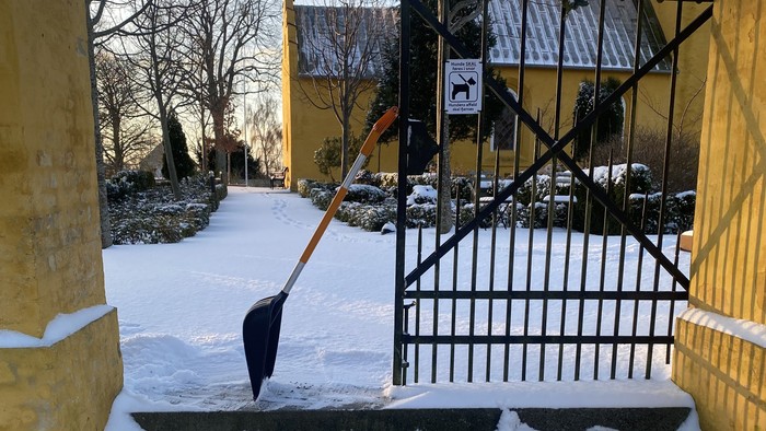Sneklædt sti med en skovl støttet op ad en bygning nær en indhegnet indgang.