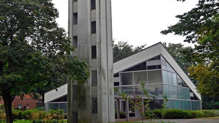 Das Bild zeigt eine moderne Kirche mit hohem Turm und Glasfront, umgeben von Bäumen.