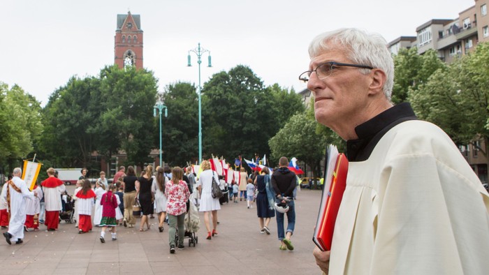Ein Mann in liturgischer Kleidung führt eine Prozession an, bei der Teilnehmer Fahnen auf einem öffentlichen Platz halten.