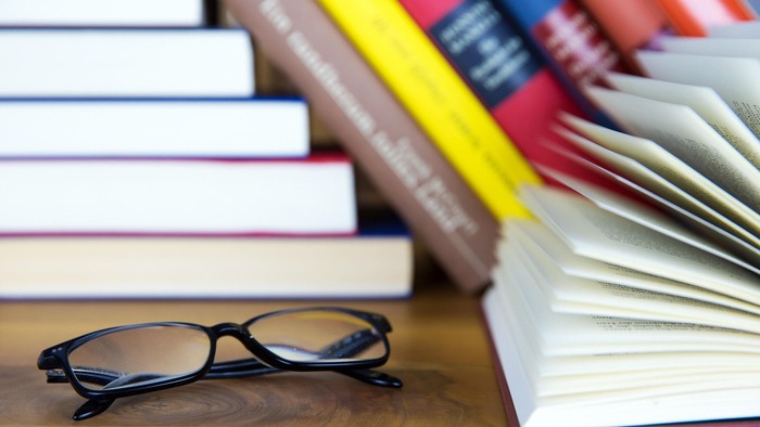 Eine Brille liegt neben einem offenen Buch und einem Stapel bunter Lehrbücher auf einem Schreibtisch.