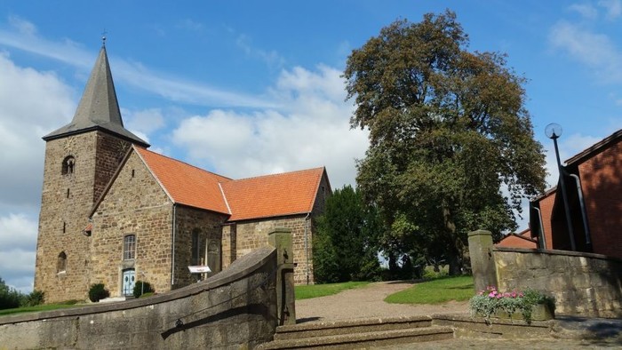 Eine mittelalterliche Kirche auf einem kleinen Hügel, rechts ein moderneres Gebäude 
