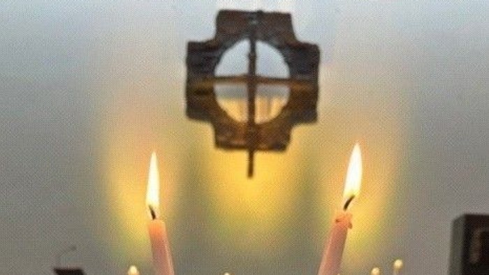 Ein ruhiger Altar zeigt drei hohe, brennende Kerzen vor einem Kreuz, umgeben von kleineren Kerzen und Blumenarrangements.