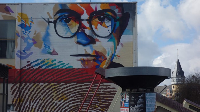 Lebendiges Wandgemälde eines stilisierten Gesichts mit bunten Mustern, neben urbanen Strukturen und einem Uhrturm.