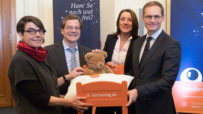 Vier Personen präsentieren ein Stofftier auf einem kleinen Bett, mit einem Schild