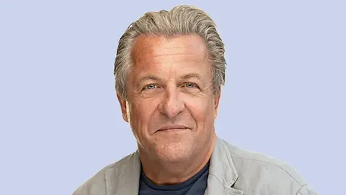 En smilende mand med gråt hår iført en lys jakke på en ensfarvet baggrund.