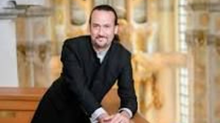 Ein Mann in eleganter Abendgarderobe sitzt anmutig auf einer Holzbank in einem prunkvollen Innenraum.