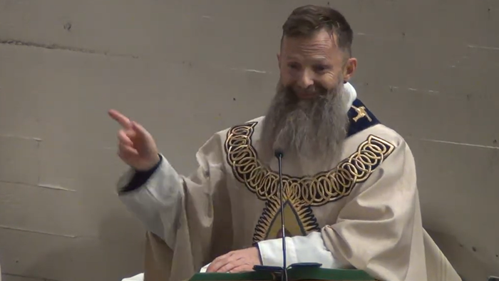 Ein bärtiger Mann in liturgischen Gewändern spricht oder predigt an einem Pult.