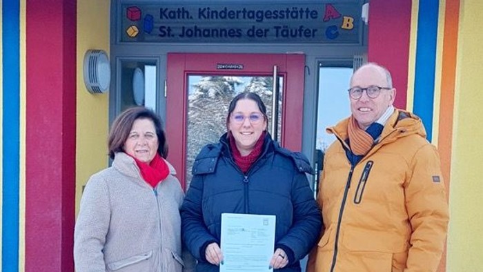 Drei Personen stehen vor einem bunten Kindergarten und halten eine Urkunde vor einem verschneiten Eingang.