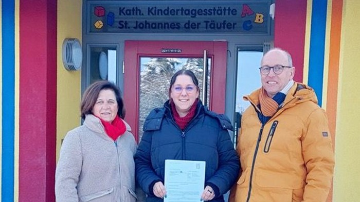 Drei Personen stehen vor einem bunten Kindergarten und halten eine Urkunde vor einem verschneiten Eingang.