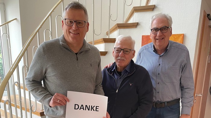 Drei Männer stehen gemeinsam drinnen und halten ein Schild mit der Aufschrift „DANKE“ als Dankesbekundung in der Nähe einer Treppe.
