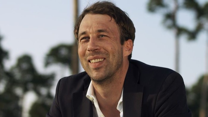 Ein lächelnder Mann im Anzug posiert im Freien zwischen Palmen.