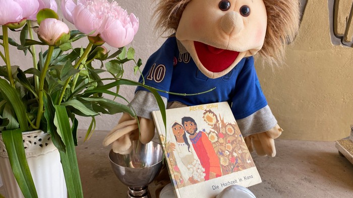 Eine Puppe hält ein Buch und sitzt neben einer Vase mit rosa Blumen.