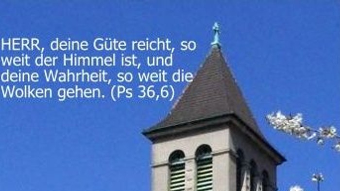Ein Kirchturm mit blühenden Bäumen und einem biblischen Zitat auf Deutsch vor einem klaren blauen Himmel.