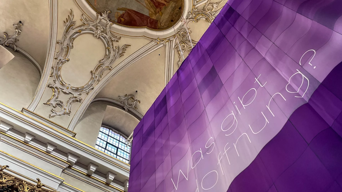 Lila Banner mit deutschem Text hängt in einem verzierten, historischen Innenarchitekturraum.