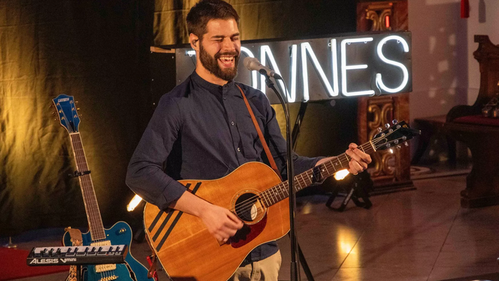 Ein Mann spielt auf der Bühne eine akustische Gitarre, während im Hintergrund eine Neonreklame mit der Aufschrift „JANNES“ leuchtet.
