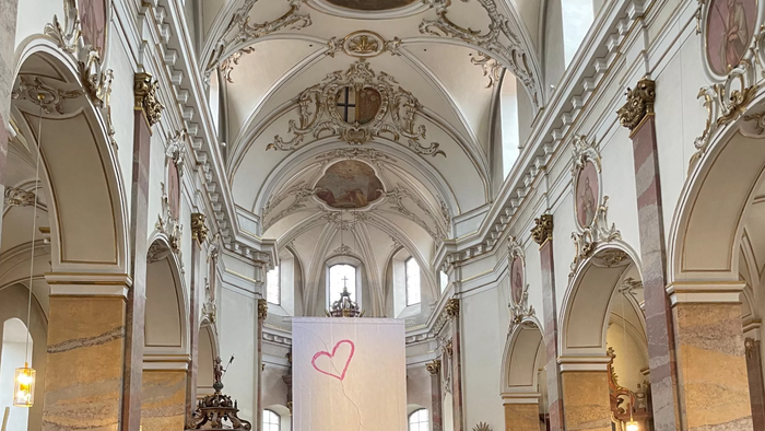 Eine überfüllte Kircheninnenraum mit prunkvollen Verzierungen, Menschen, die an einem Gottesdienst teilnehmen, und ein Herz-Banner auf dem Altar.