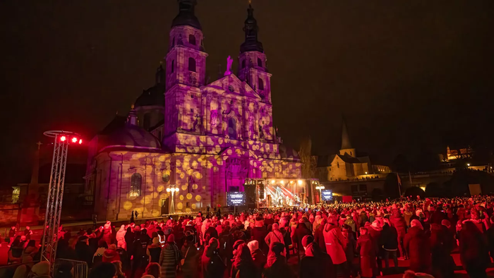 Lebendiges Nachtfest mit beleuchteter Kathedrale und großer Menschenmenge, die im Freien feiert.