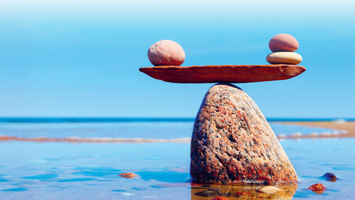 Ein roter Granitfelsen ragt aus dem Meer, darüber eine weitere Steinplatte, auf beiden Seiten sind kleinere Steine geschichtet, wie auf einer Waage.