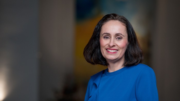 Professionelle Frau in einem blauen Blazer, die selbstbewusst vor einem modernen Innenhintergrund lächelt.