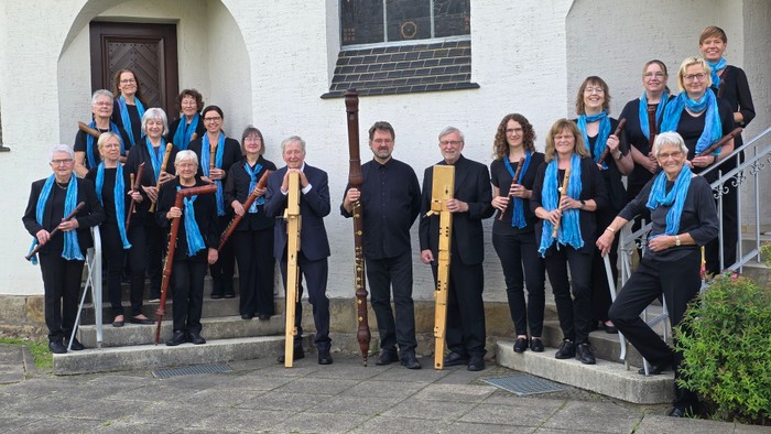 Eine Gruppe von Musiker:innen in formeller Kleidung mit blauen Schals, die draußen vor einem Gebäude posieren und verschiedene Blasinstrumente halten.
