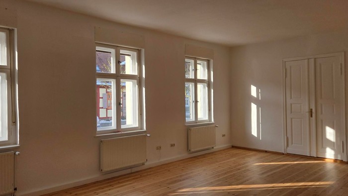 Heller, leerer Raum mit großen Fenstern, Holzböden und Heizkörpern unter den Fensterflügeln.