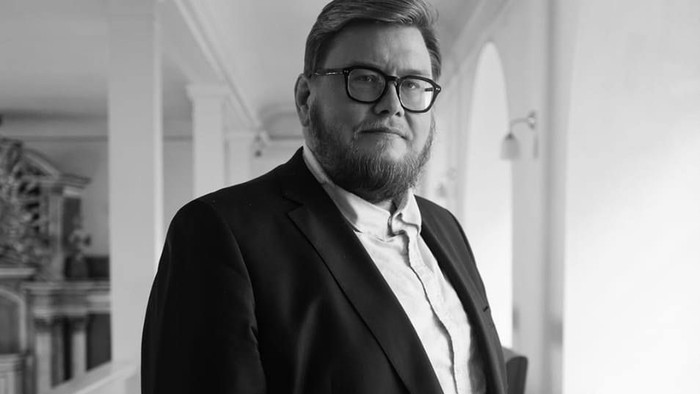 En mand i jakkesæt og briller står selvsikkert i en veloplyst indendørs gang.