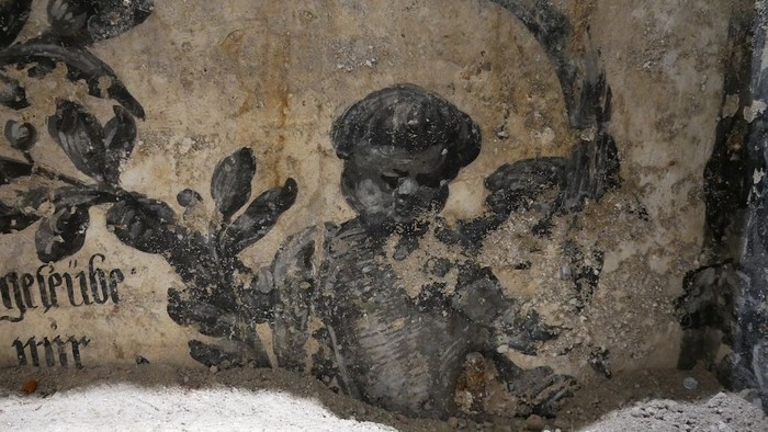 Alte Wandmalerei, die eine skelettartige Figur zeigt, die ein Kind mit Heiligenschein inmitten von gealtertem, rissigem Putz hält.