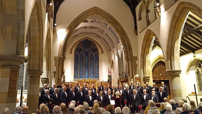 A choir performs in a grand, historic church with an attentive audience seated in red chairs.