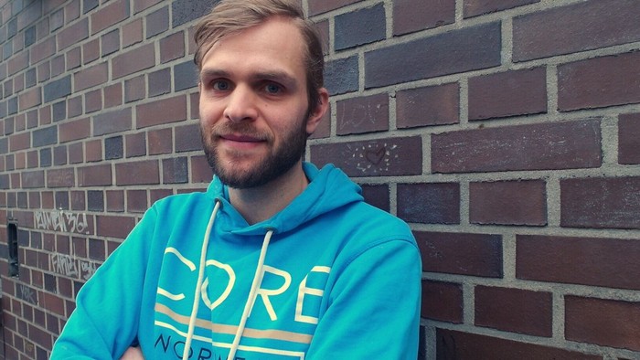 Ein Mann mit blonden Haaren und Bart steht vor einer Ziegelmauer und trägt ein blaues Kapuzenpullover mit gelbem Text.