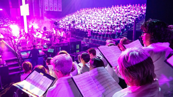 Eine lebhafte Chorprobe unter farbenfroher Bühnenbeleuchtung mit Musiker:innen und Publikum im Hintergrund.