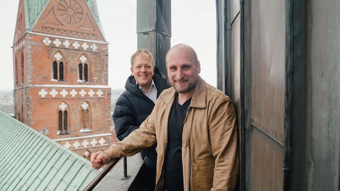 Zwei Personen, die auf einem Balkon Händchen halten, mit einer historischen Kirche im Hintergrund.