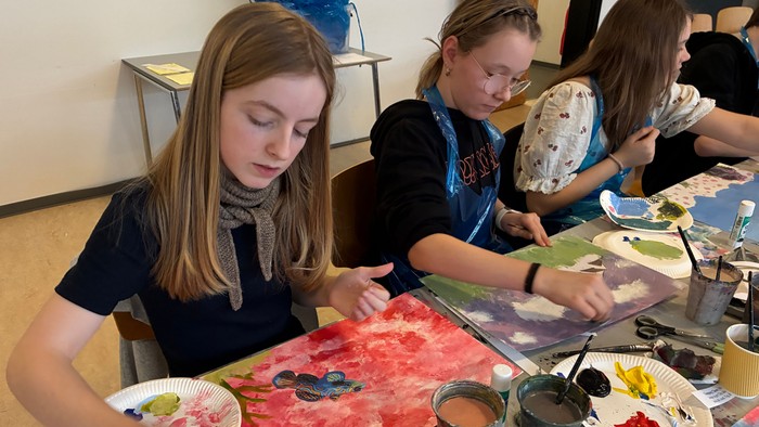 Unge mennesker maler sammen på store lærreder med forskellige kunstmaterialer ved et bord.